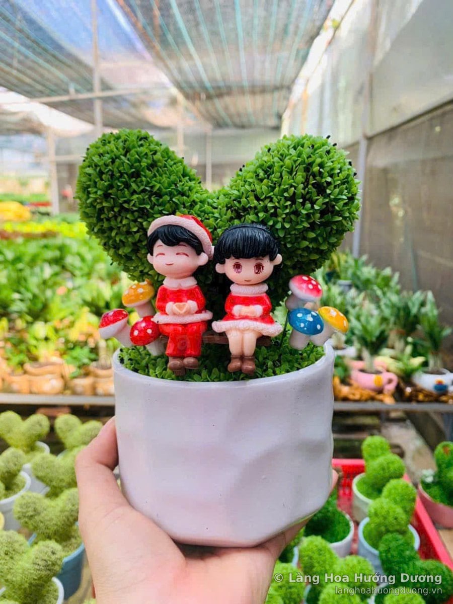 Chậu sứ trắng nhỏ có cụm cây cắt tỉa hình tim, hai tượng nhỏ mặc đồ Noel ngồi trên ghế và vài cây nấm trang trí